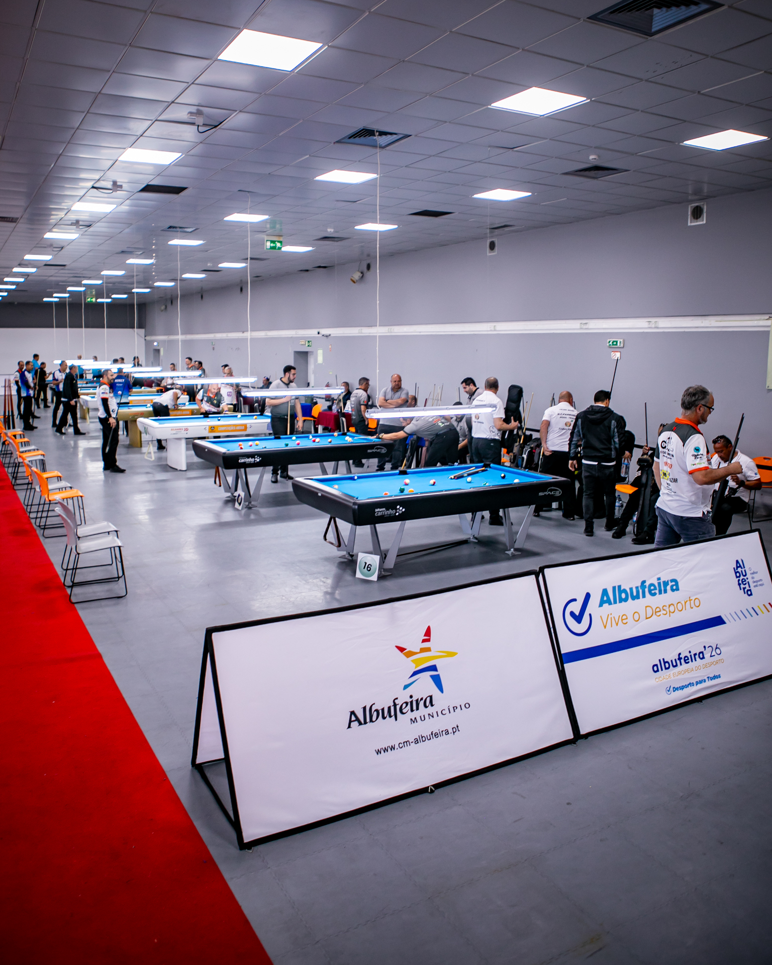 Sala de competição com mesas de bilhar, jogadores ao redor e placas de torneio, sob luzes do teto.