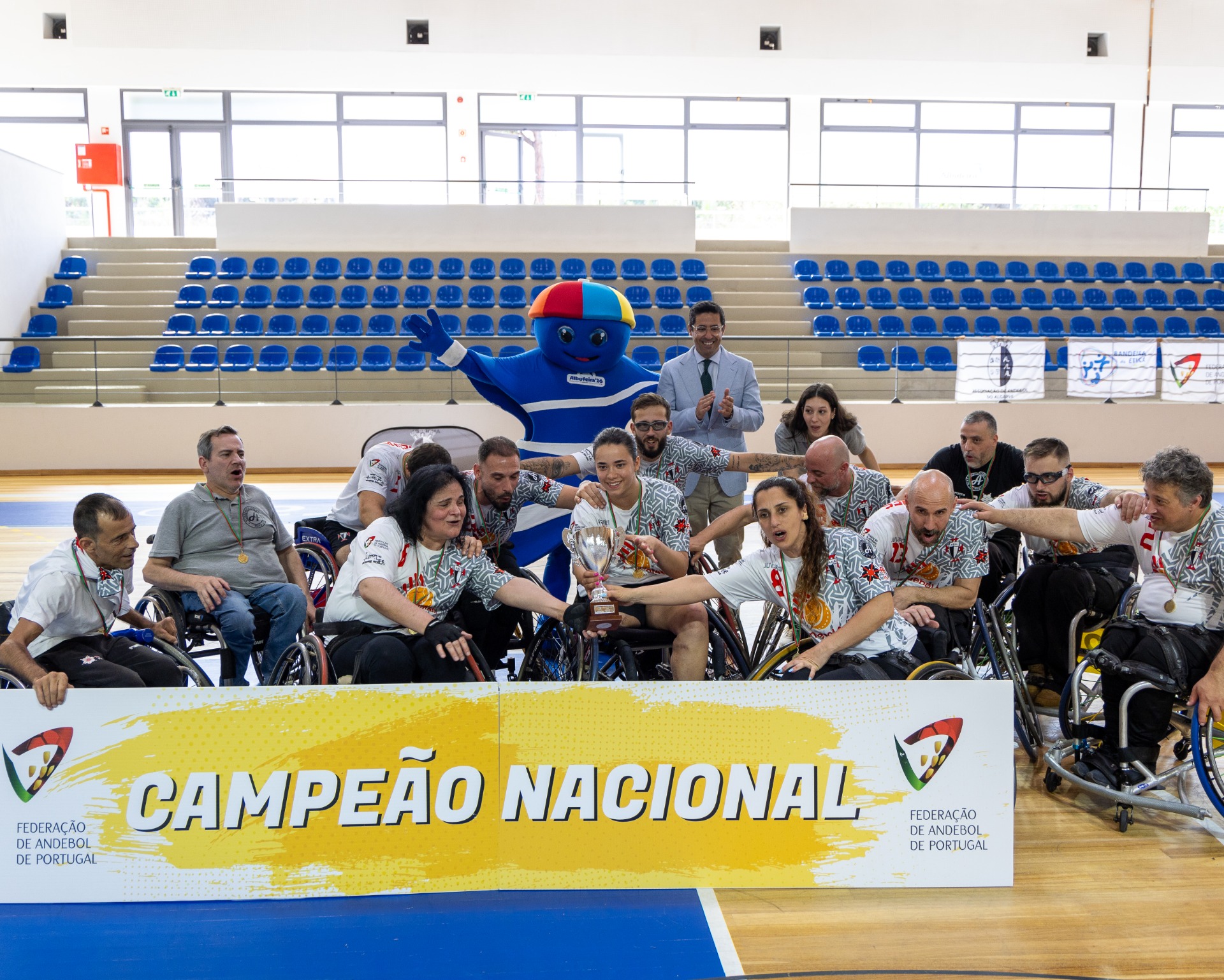 Equipa de andebol em cadeiras de rodas celebra, com troféu, mascote azul e banner “Campeão Nacional”.