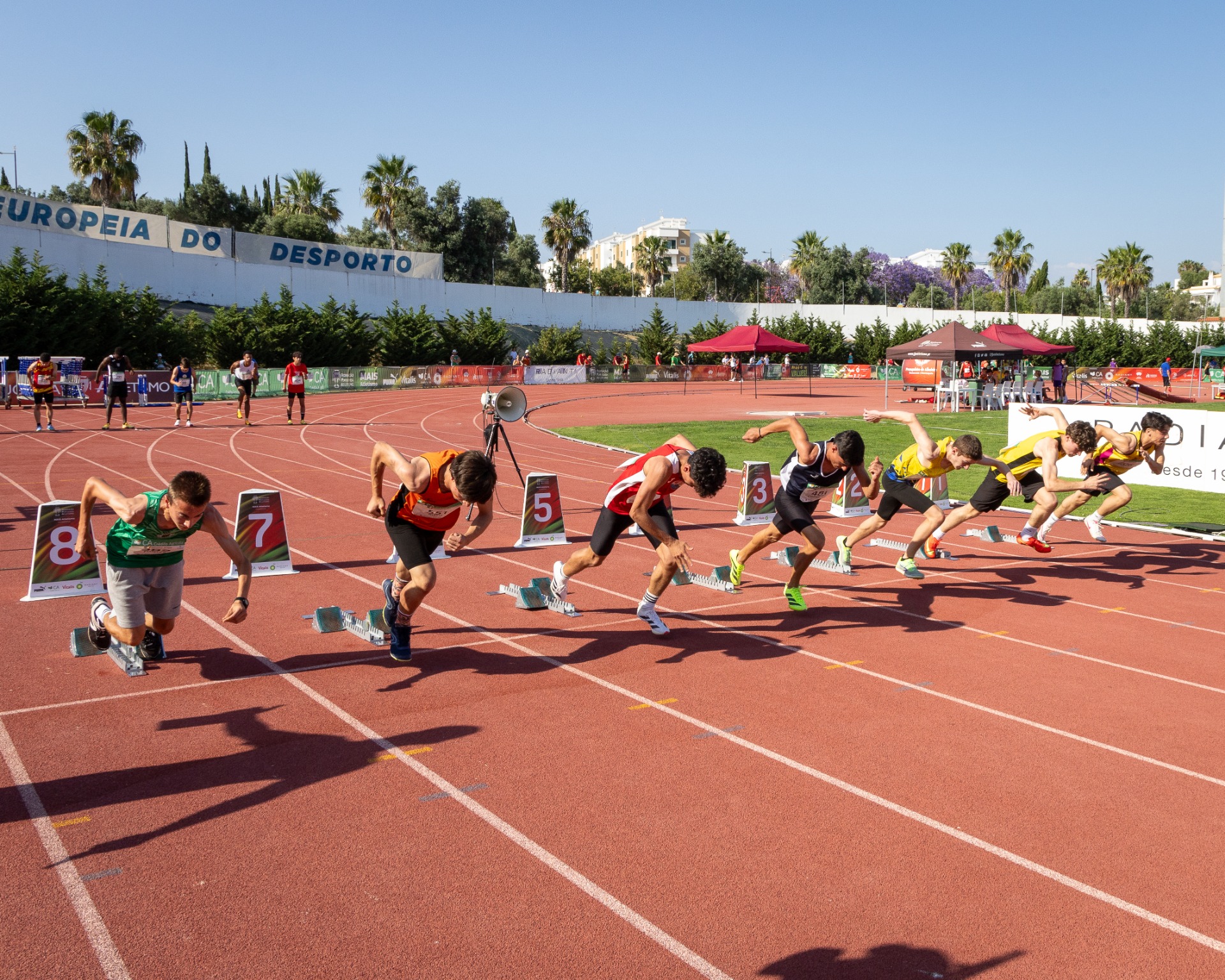Atletas em corrida de pista vermelha, partindo de blocos, com tendas e palmeiras ao fundo.