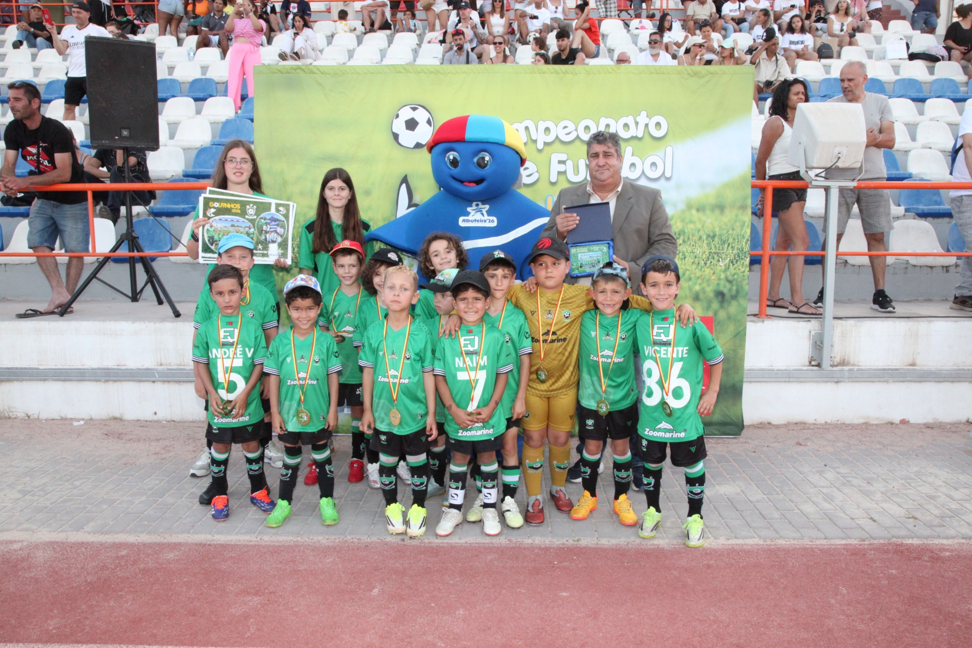 Grupo de crianças e treinador posam com troféus no estádio, ao lado de mascote azul.
