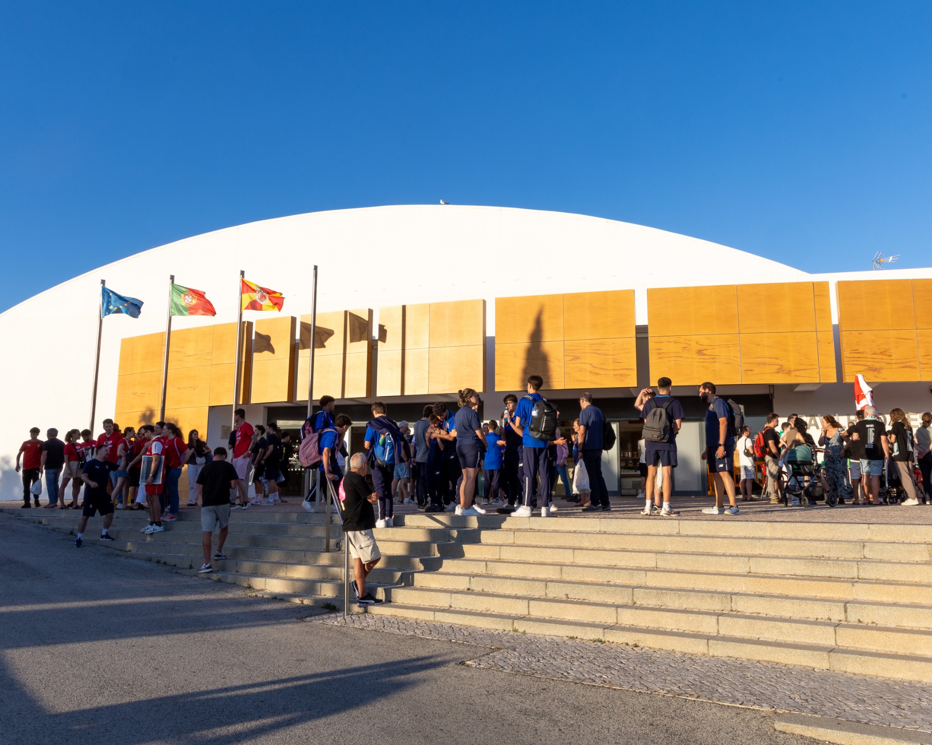 Grupo de pessoas em fila diante de um grande pavilhão branco com bandeiras ao céu azul.