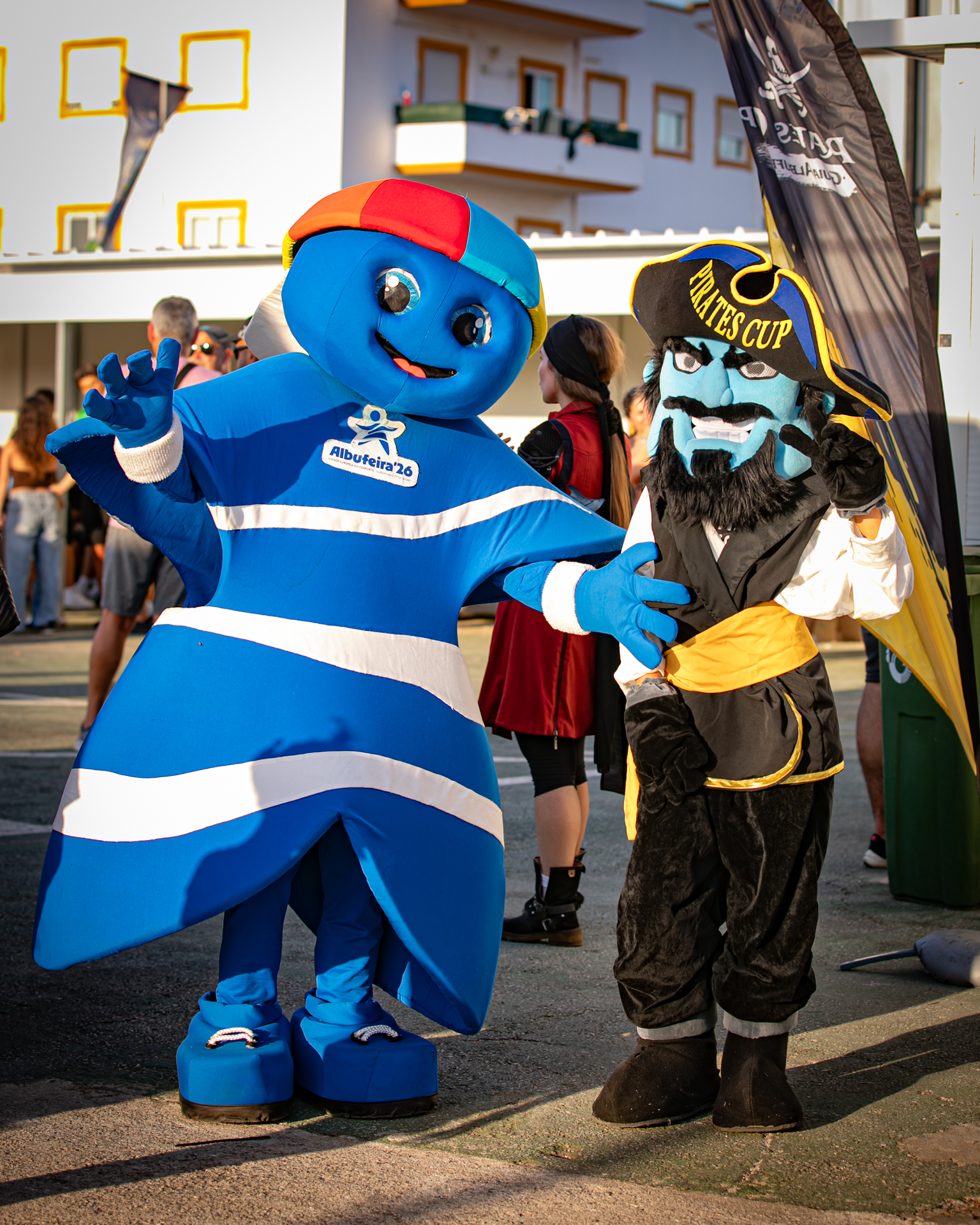 Personagens mascote azuis e pretos cumprimentam crianças em evento ao ar livre, com fachada ao fundo.