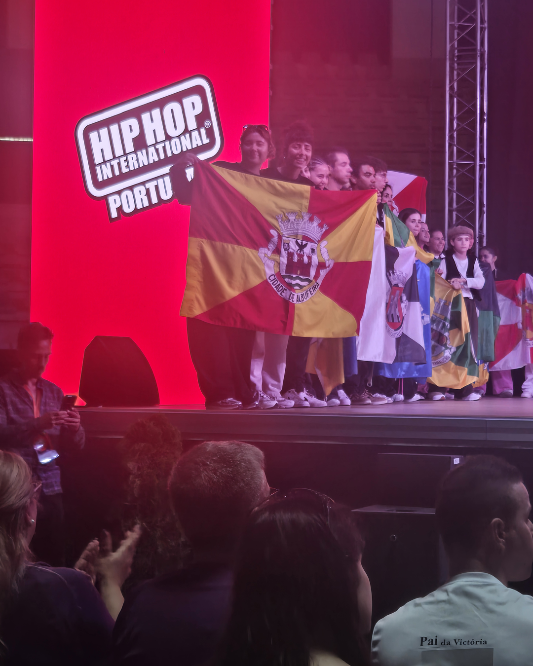 Palco de evento com bandeira, pessoas cantando e público ao fundo sob luzes vermelhas.