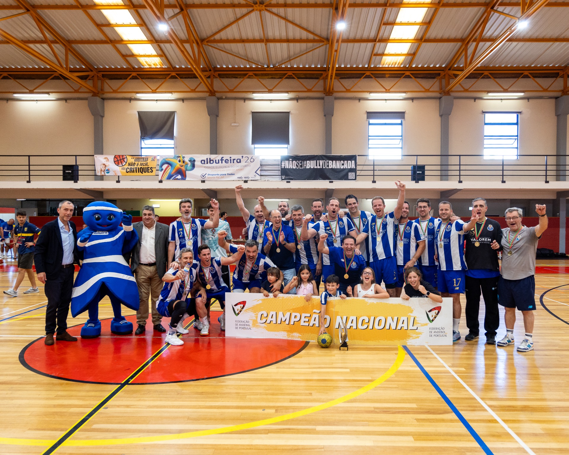 Equipa de andebol celebra vitória em pavilhão, com mascote azul e banner “Campeão Nacional”.