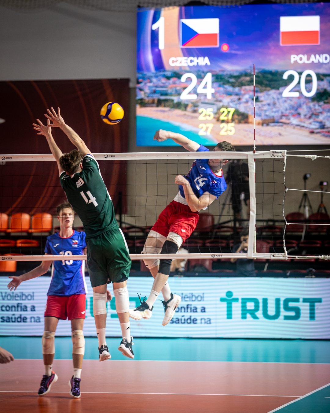 Jogadores de voleibol em campo, um salta para bloquear a bola sobre a rede.