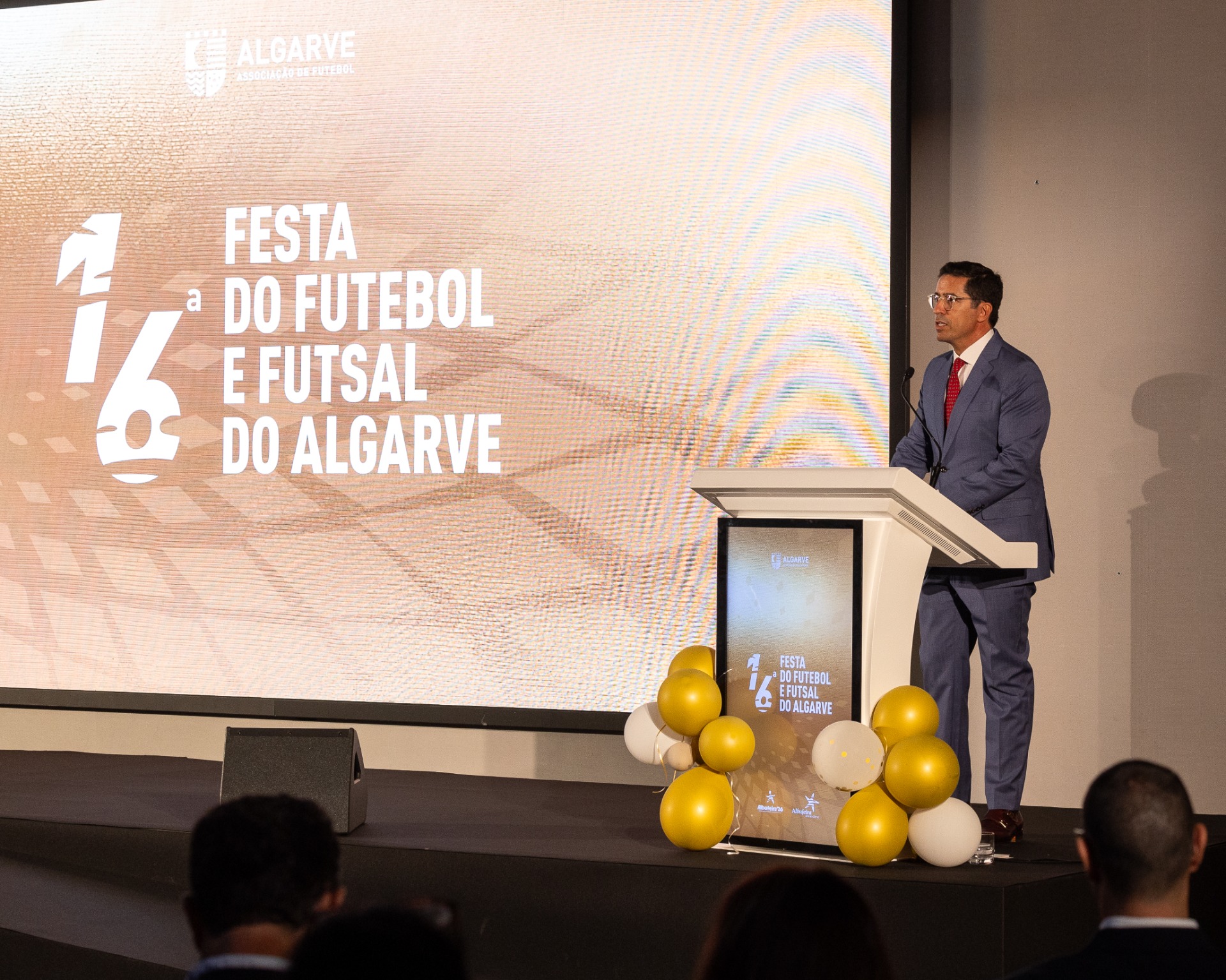 Homem em palco com fato, a discursar num pódio, balões dourados e ecrã com “Festa do Futebol”.