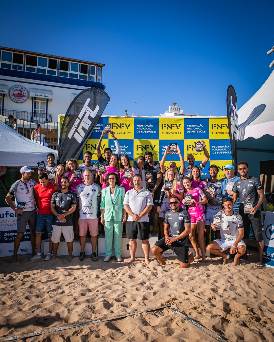 Grupo de atletas posa na areia diante de bandeiras e banner da Federação Nacional de Futevólei.