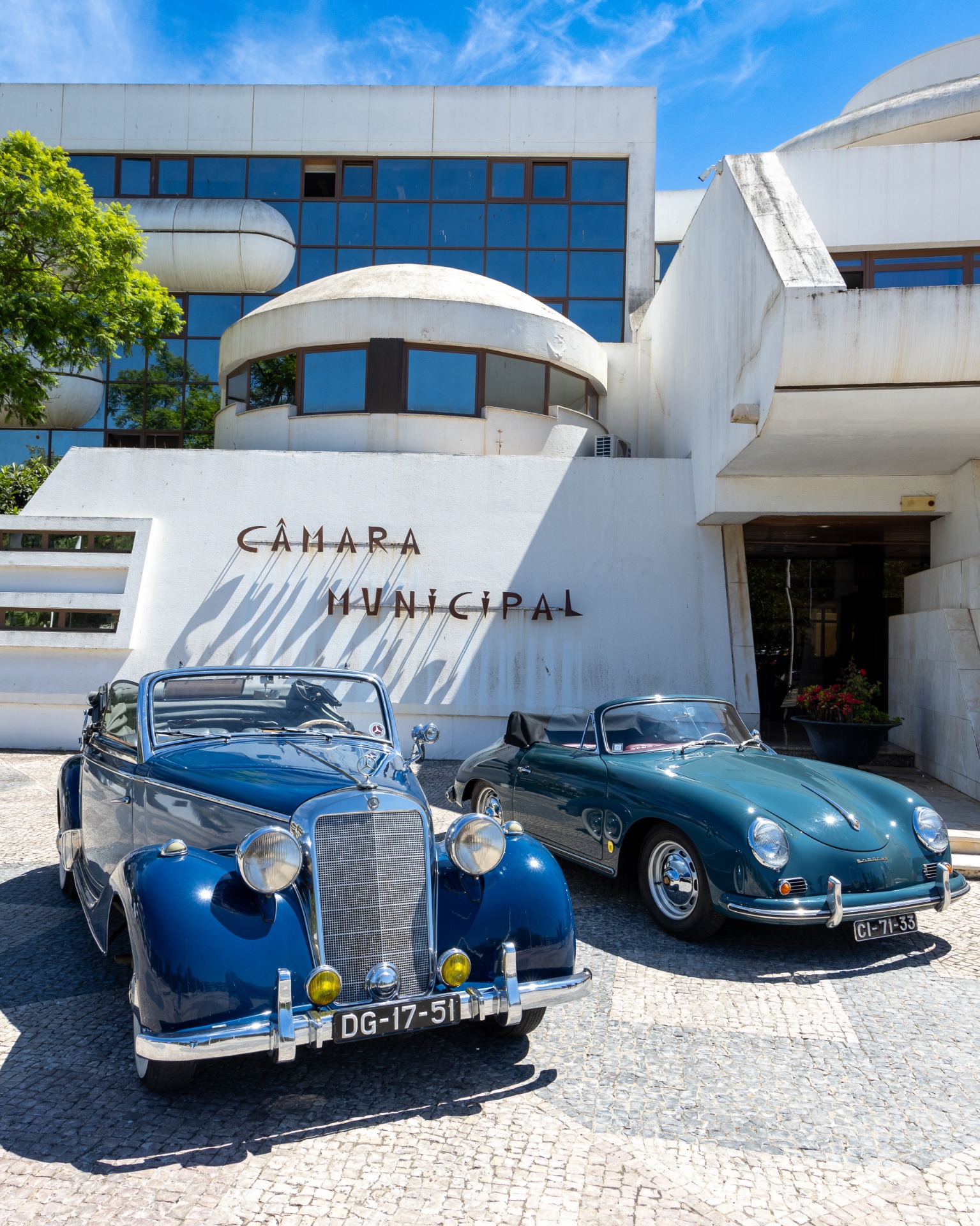 Dois carros clássicos azuis estacionados em frente ao prédio branco da Câmara Municipal, com céu azul.