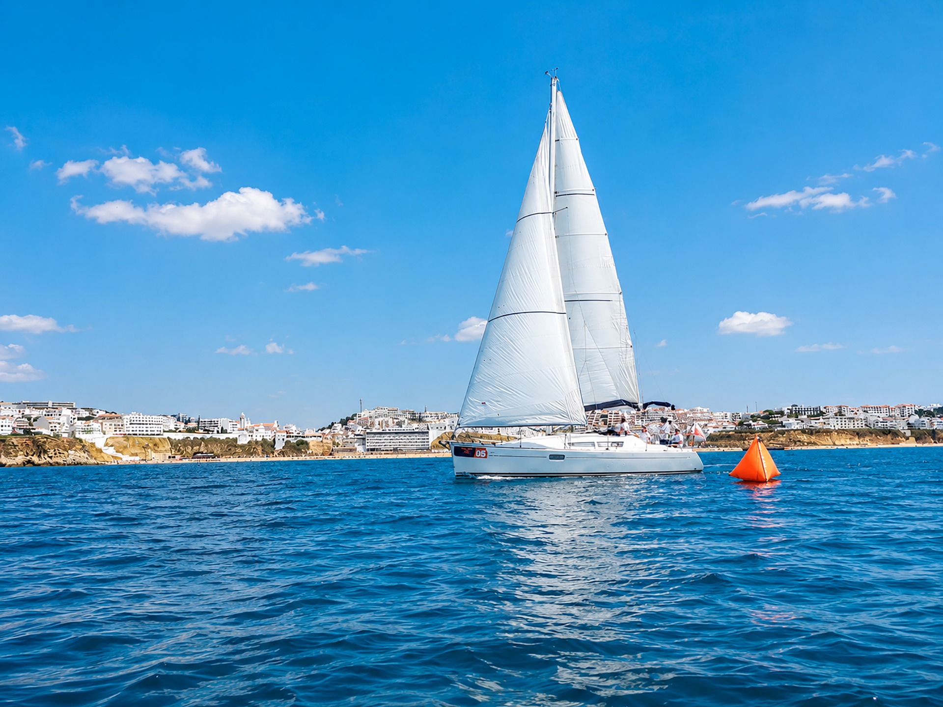 Iate à vela branco navegando no mar azul, com vela aberta e cidade costeira ao fundo.