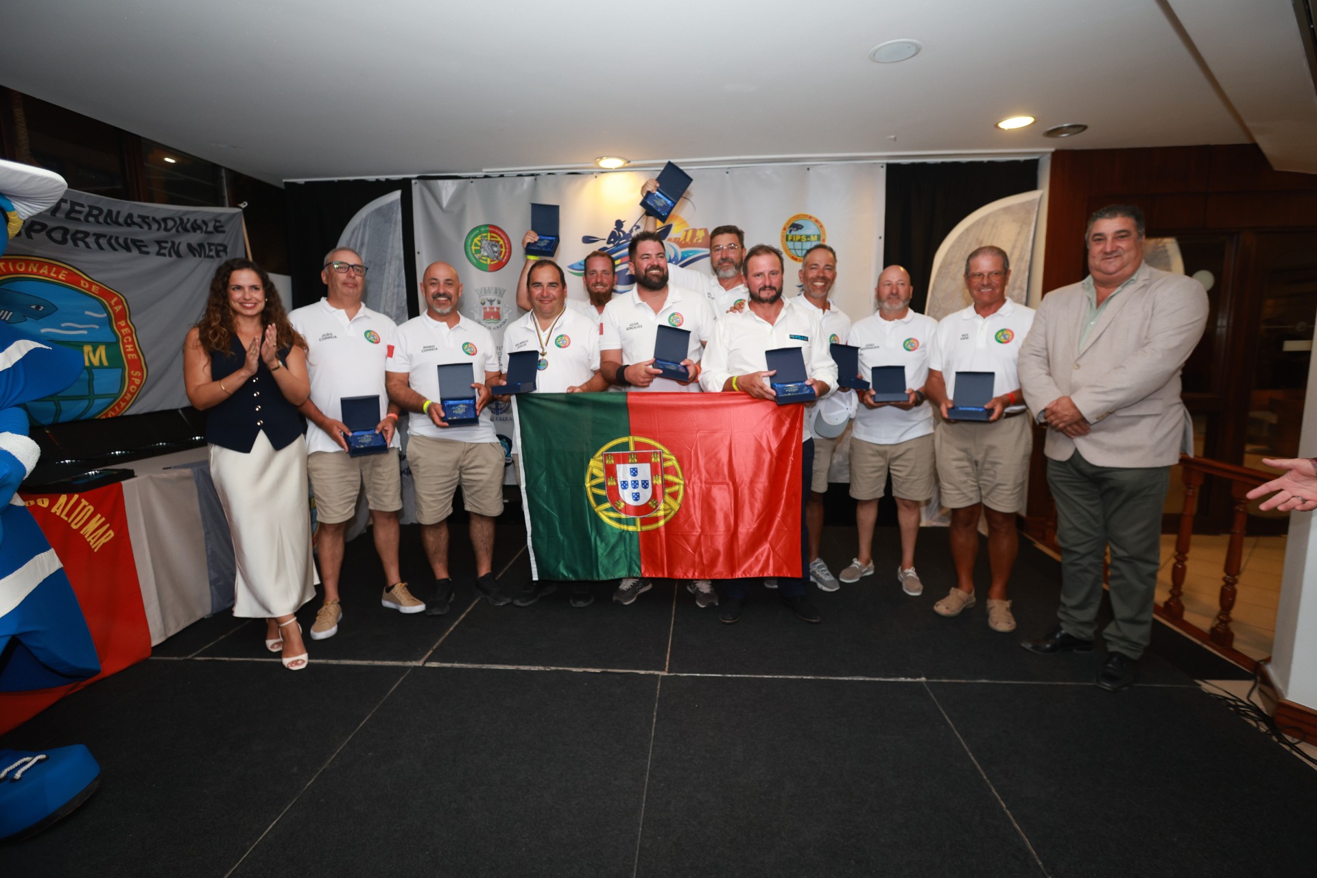 Grupo de pessoas em evento, segurando troféus e caixas, com bandeira portuguesa ao centro.