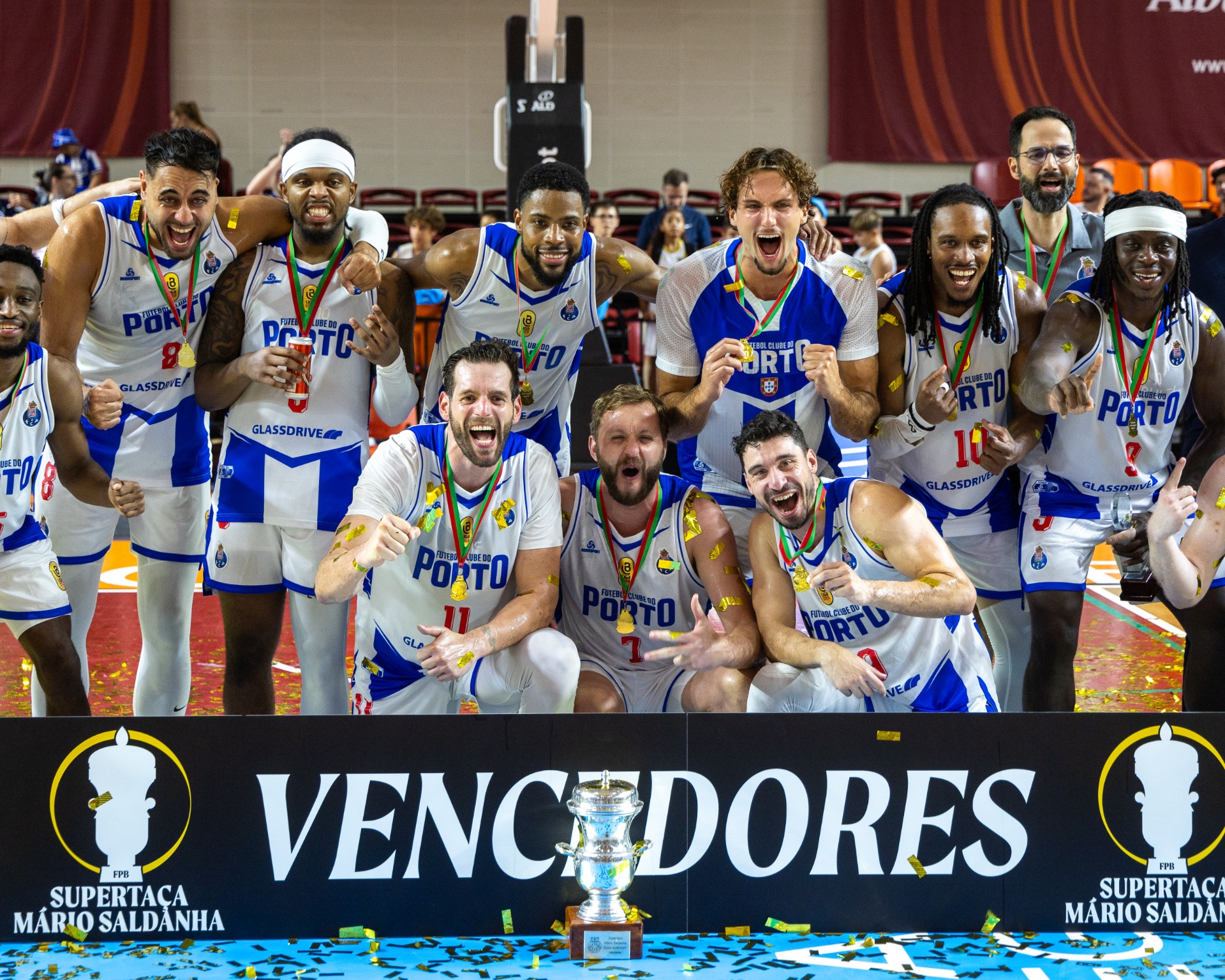 Jogadores de basquetebol comemoram com medalhas e taça, pulando e sorrindo em palco coberto.