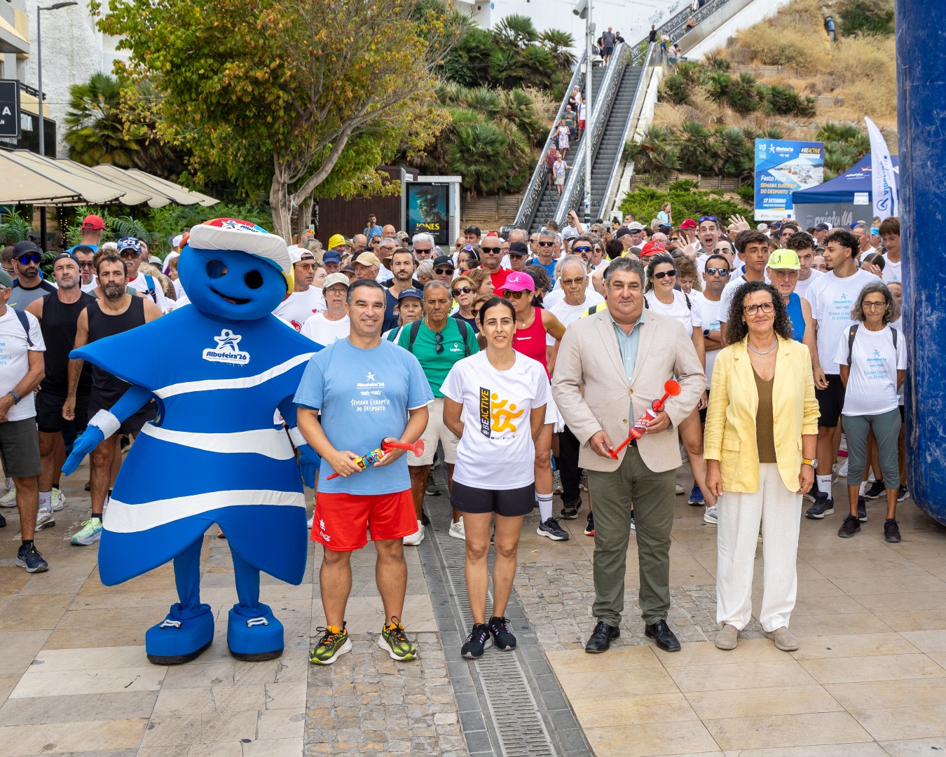 Multidão em evento desportivo com mascote azul e listras, pessoas sorrindo e escadas ao fundo.