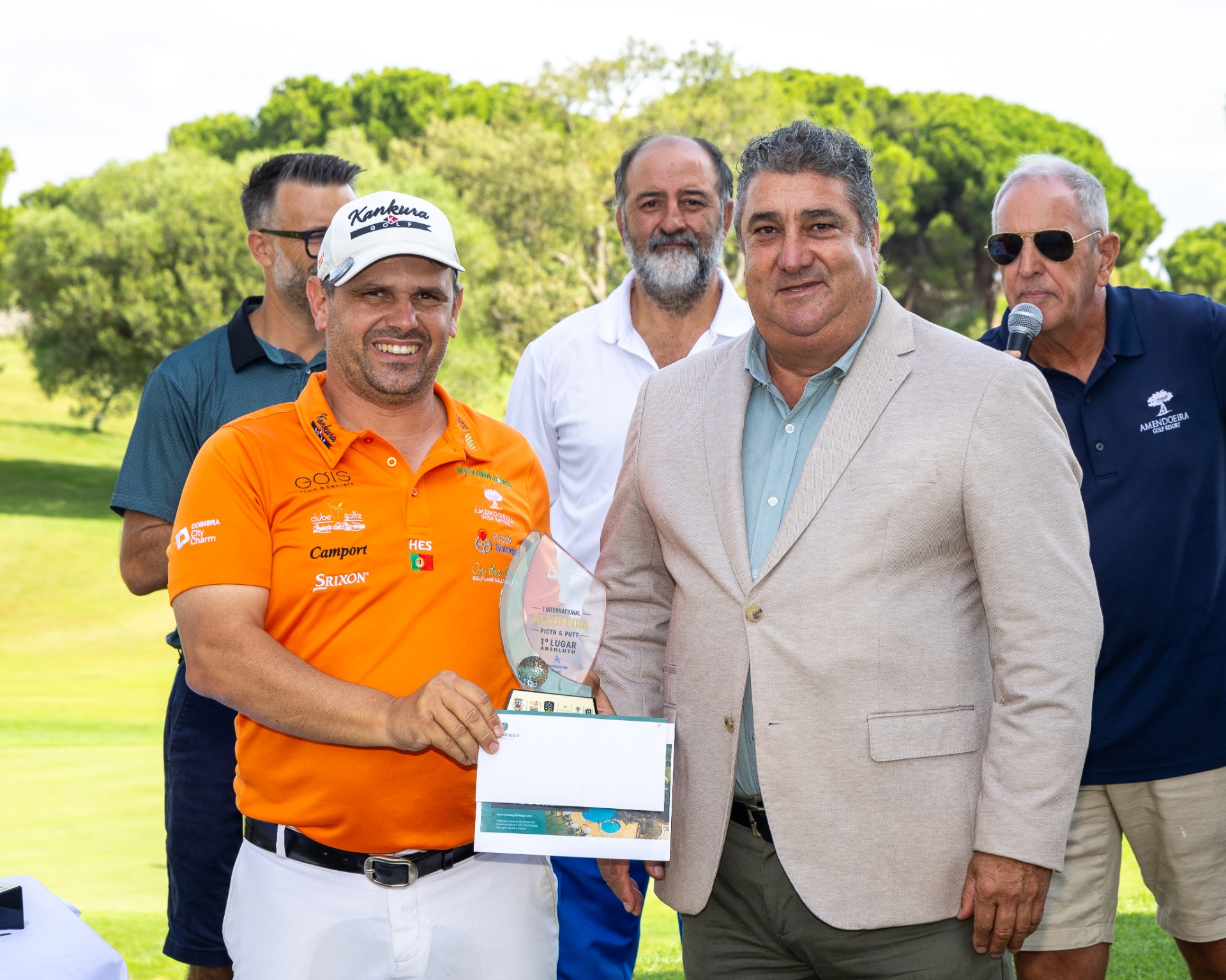 Homens em campo de golfe, um jogador de laranja segura troféu e certificado, ao lado de organizadores.