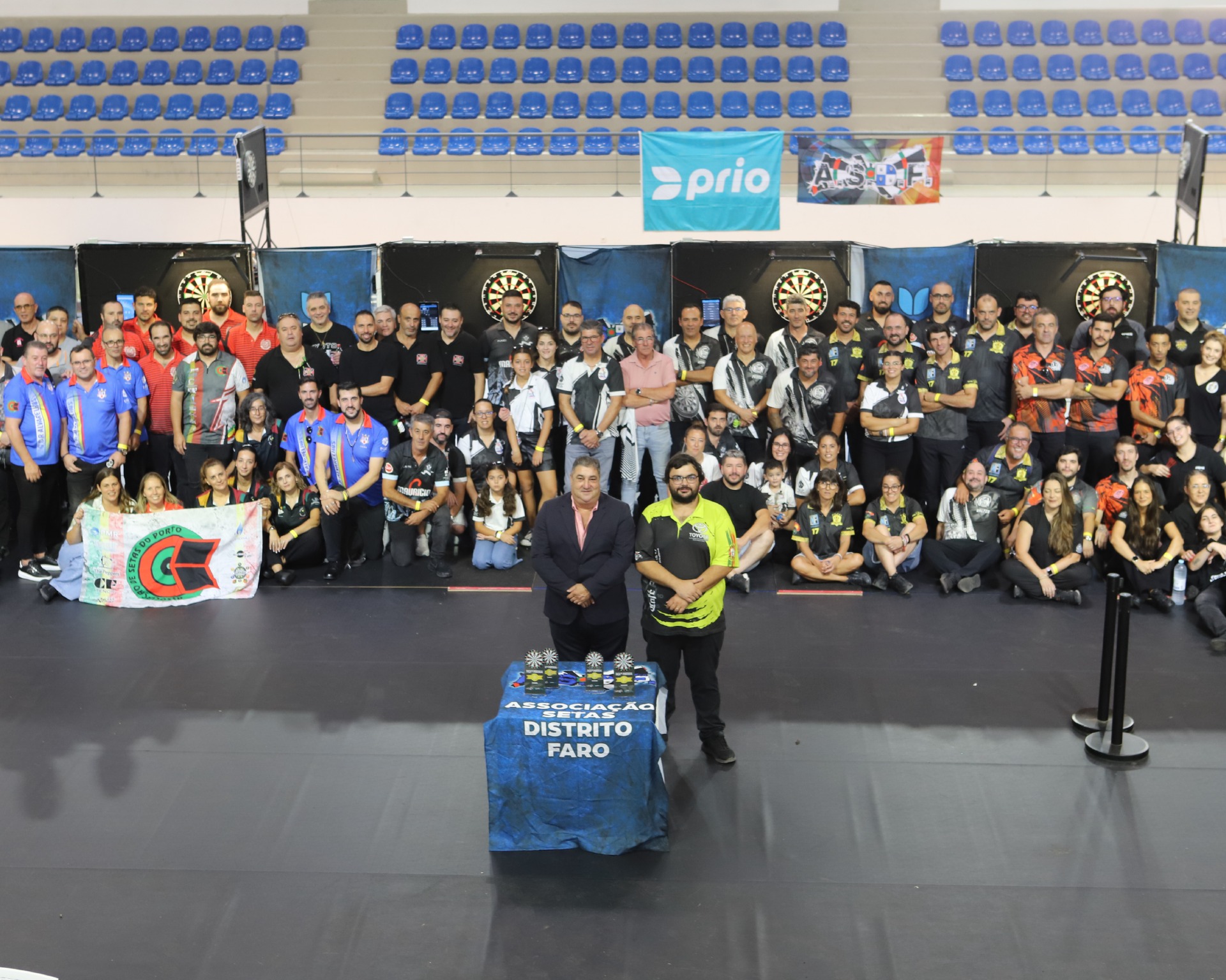 Grupo grande posa em arena de dardos, com banners, alvos e bandeiras ao fundo.