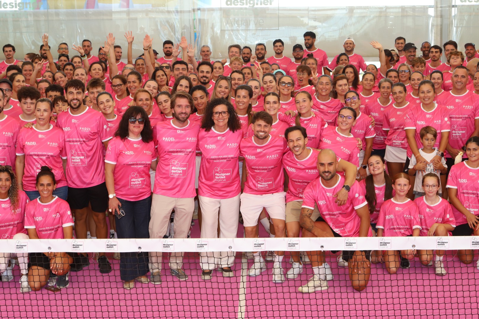 Grupo grande de pessoas sorridentes em camisolas rosa, posando juntas num evento solidário de padel.