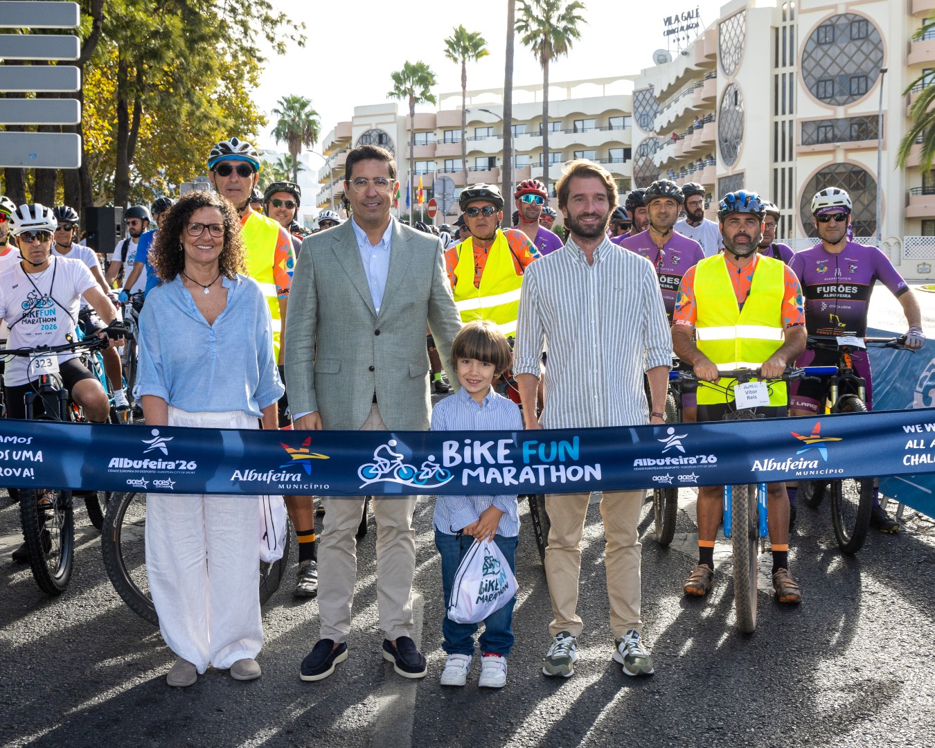 Grupo de pessoas em fila, com bicicletas, em evento Bike Fun Marathon, ao lado de prédio ensolarado.