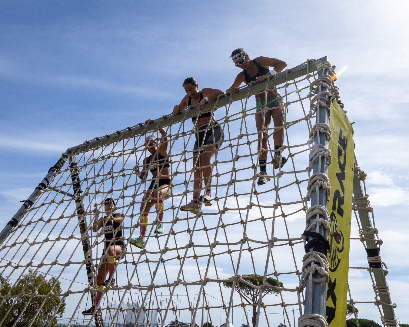 Atletas escalam uma rede de cordas alta em evento desportivo, com céu azul ao fundo.