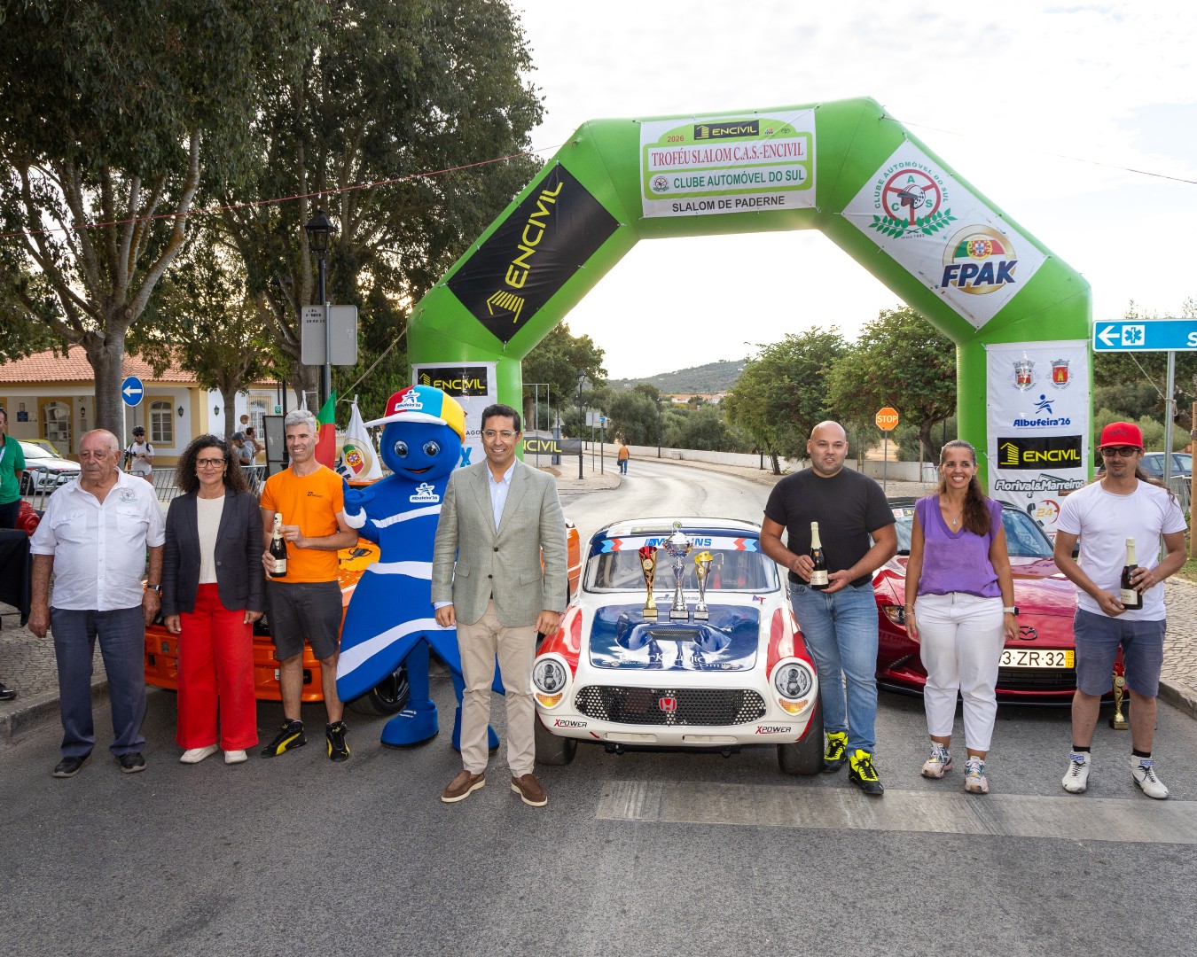Grupo de participantes posa com troféus diante de carros clássicos sob arco inflável verde de evento automóvel.
